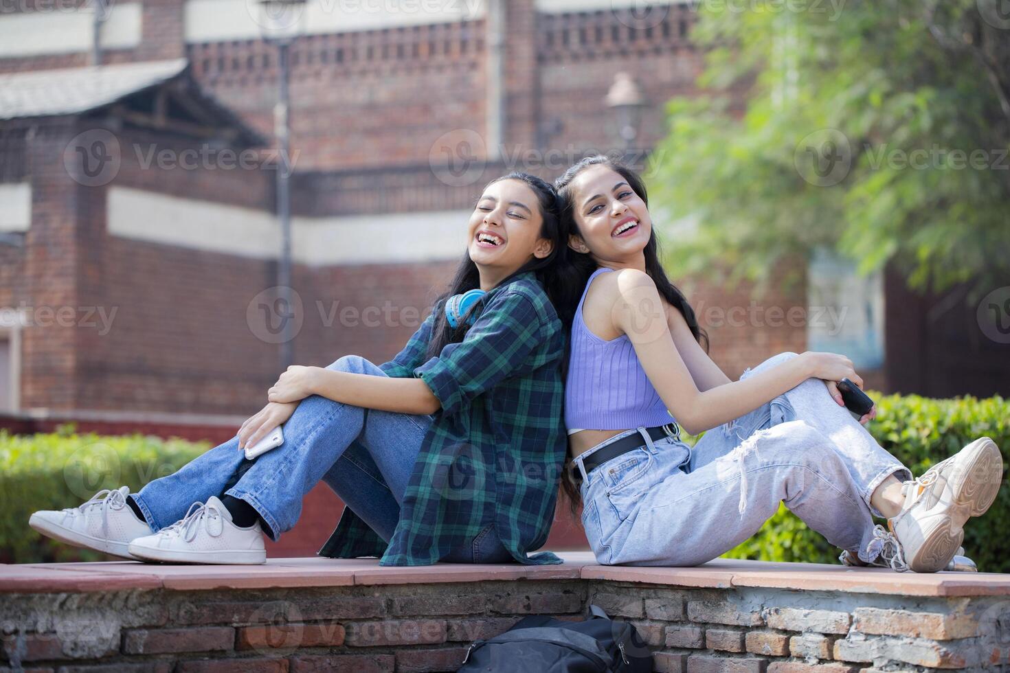 Fashion Girls Laughing While Sitting Back to Back in the Park photo