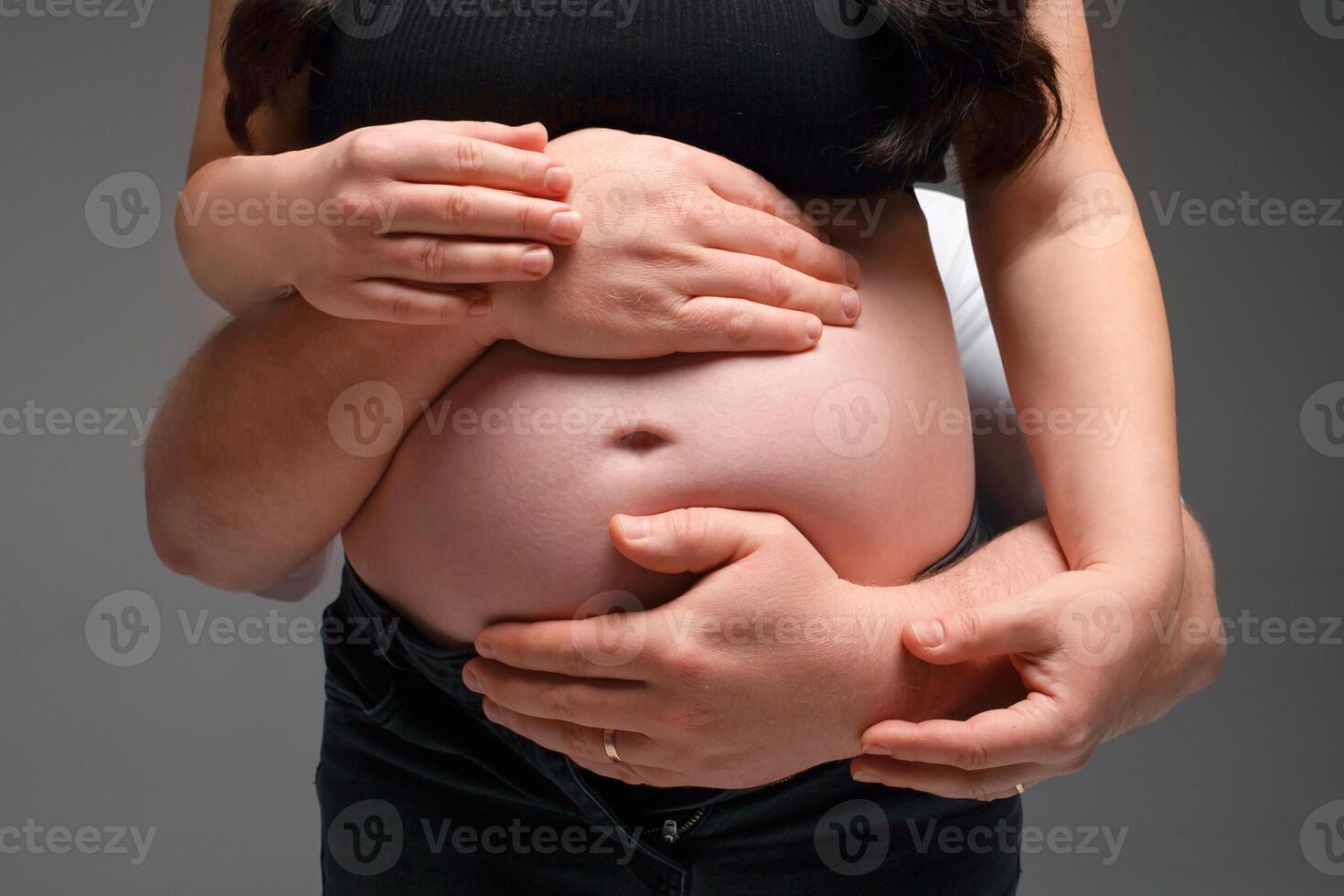 close-up photograph of a pregnant belly being hugged by the parents' hands on a gray background. family expecting a baby photo