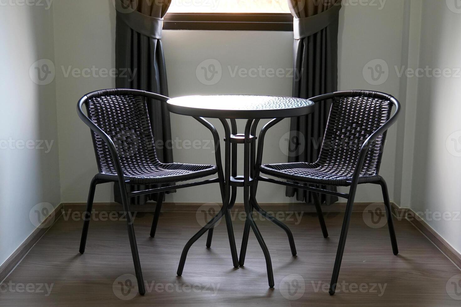 The faux rattan table and chair set consists of 2 chairs and a black round table placed next to the window with black curtains in the bedroom for sitting or working. photo
