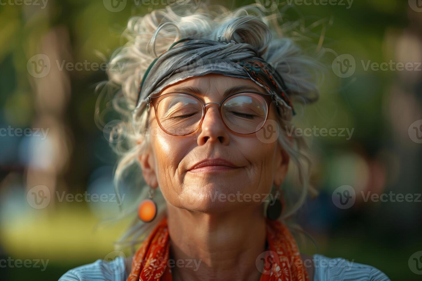 A senior woman with gray hair, wearing glasses and a headband, is practicing deep breathing exercises in a park. She has her eyes closed and a serene expression on her face photo