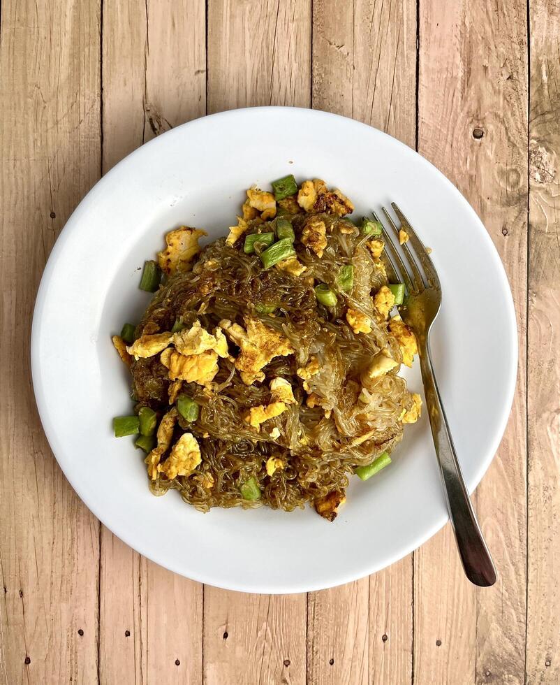 Stir fried sweet soy sauced rice noodle vermicelli with scrambled eggs and long beans in plate from above top view angle isolated food photography on vertical wooden background. Bihun goreng telur. photo