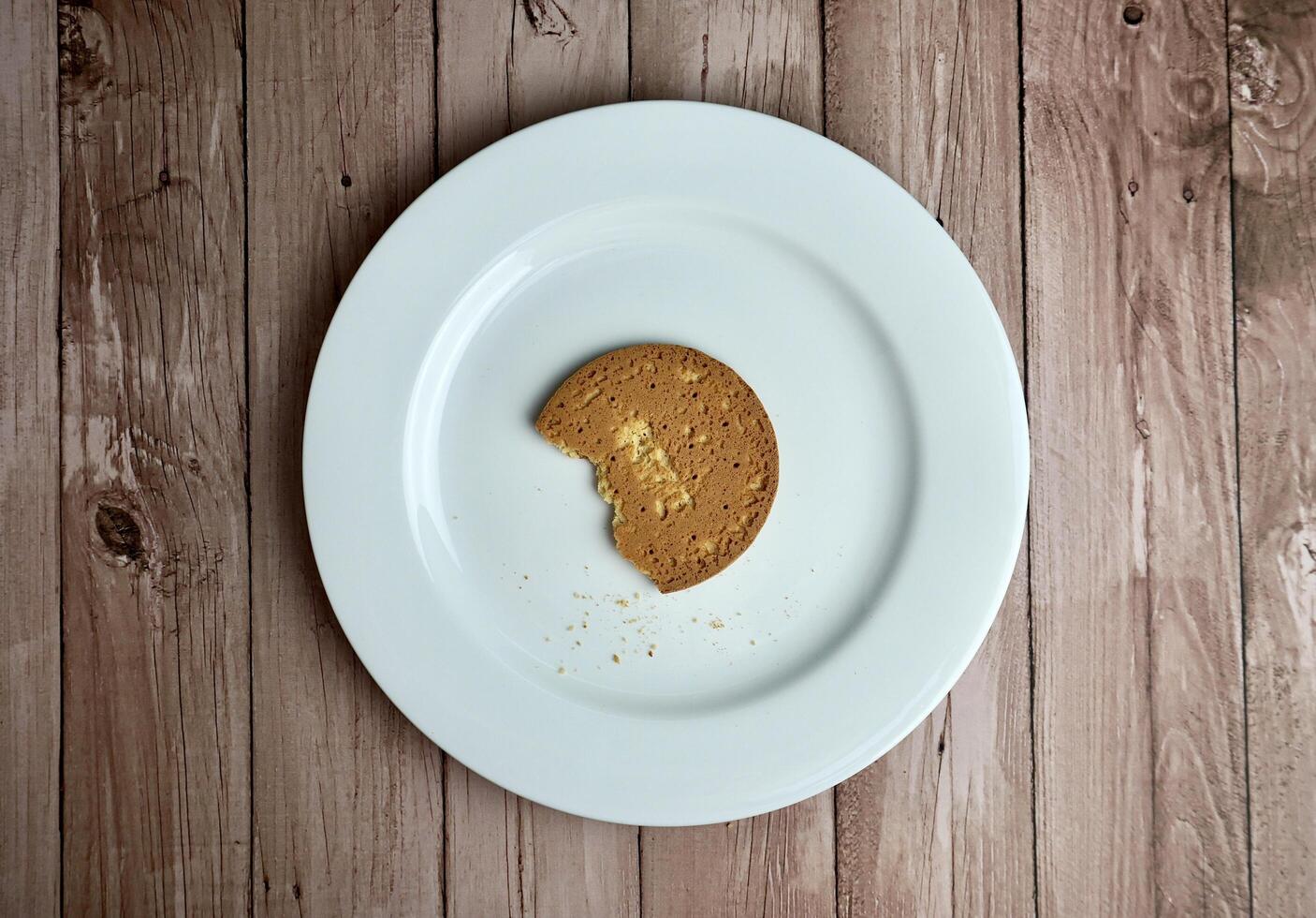 One last piece of single bitten round brown sweet sugary butter biscuit on top of white plate. Food object photography from above top view angle isolated on wooden surface background. photo