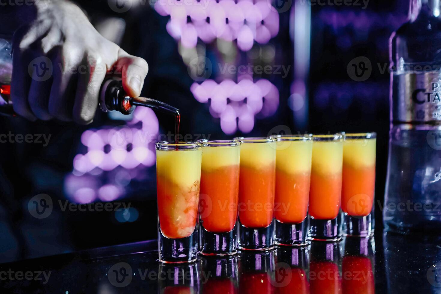 Bartender Pouring Cocktail Into Row of Shot Glasses photo