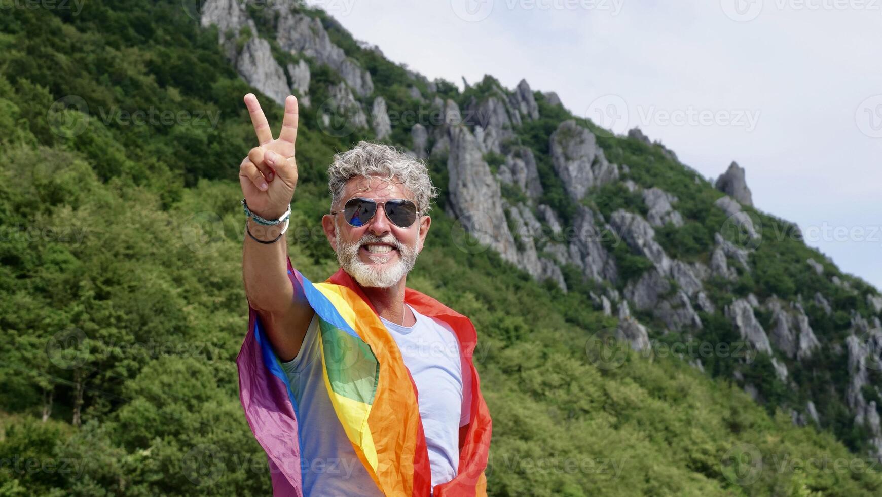 Portrait of a gray-haired elderly Caucasian man with a beard and sunglasses holding a rainbow LGBTQIA flag against a sky background, shouts in protest, Celebrates Pride Month Coming Out Day photo