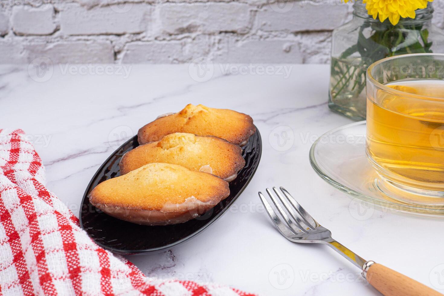 Lemon glaze madeleines on black a plate with a cup of tea placed on a marble background with a white brick wall. Madeleine - homemade traditional French small cookie. Seashell sweet cakes photo