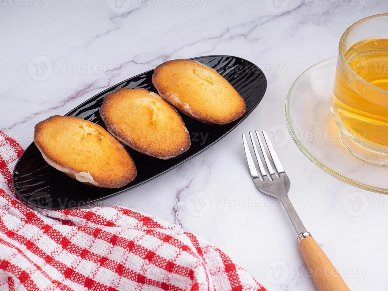 Lemon glaze madeleines on black a plate with a cup of tea placed on a marble background with a white brick wall. Madeleine - homemade traditional French small cookie. Seashell sweet cakes photo