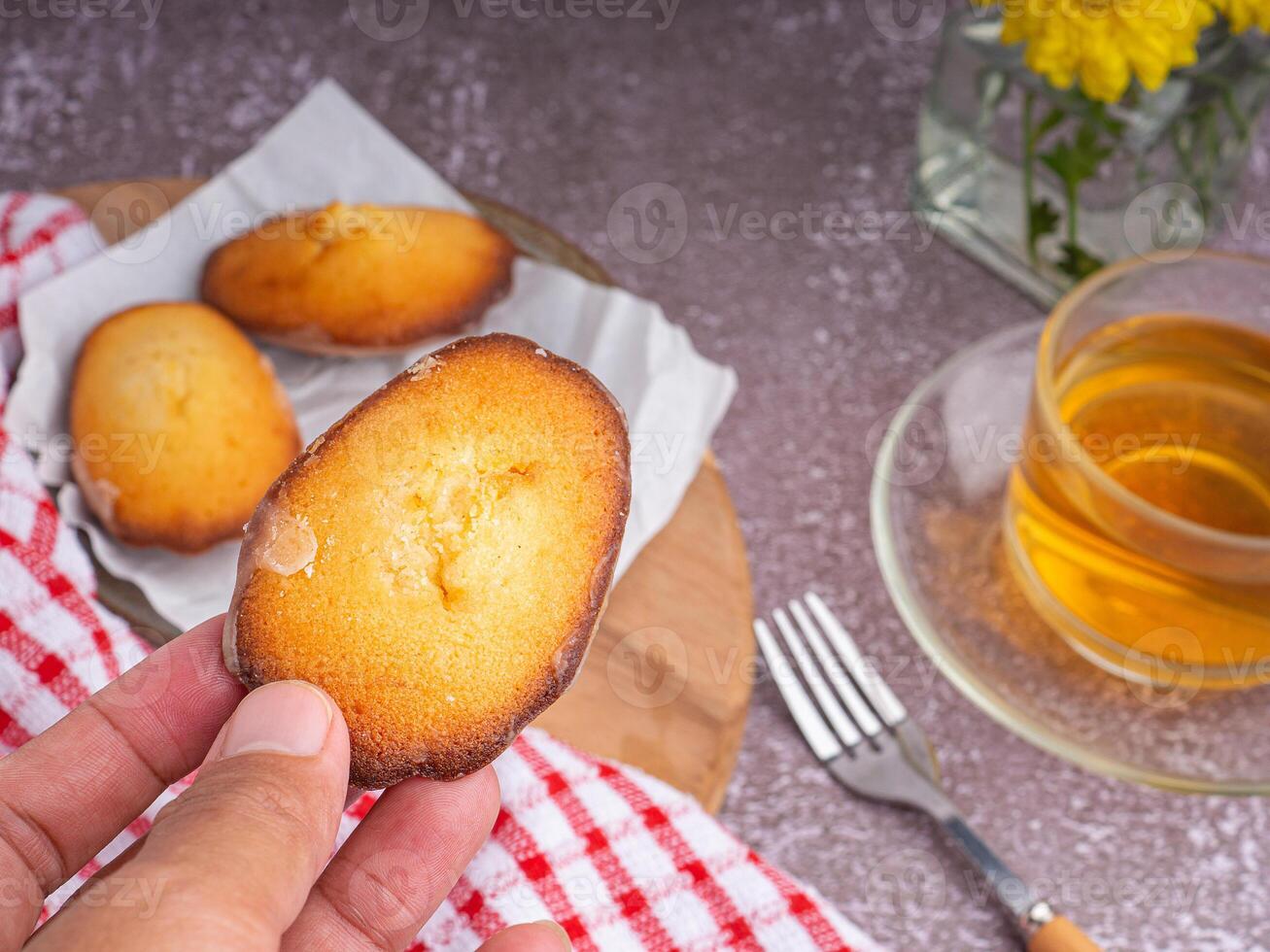 Hand holding a piece of madeleine on lemon glaze madeleines on a plate with a cup of tea placed on a gray stone background. Madeleine - homemade traditional French small cookie photo