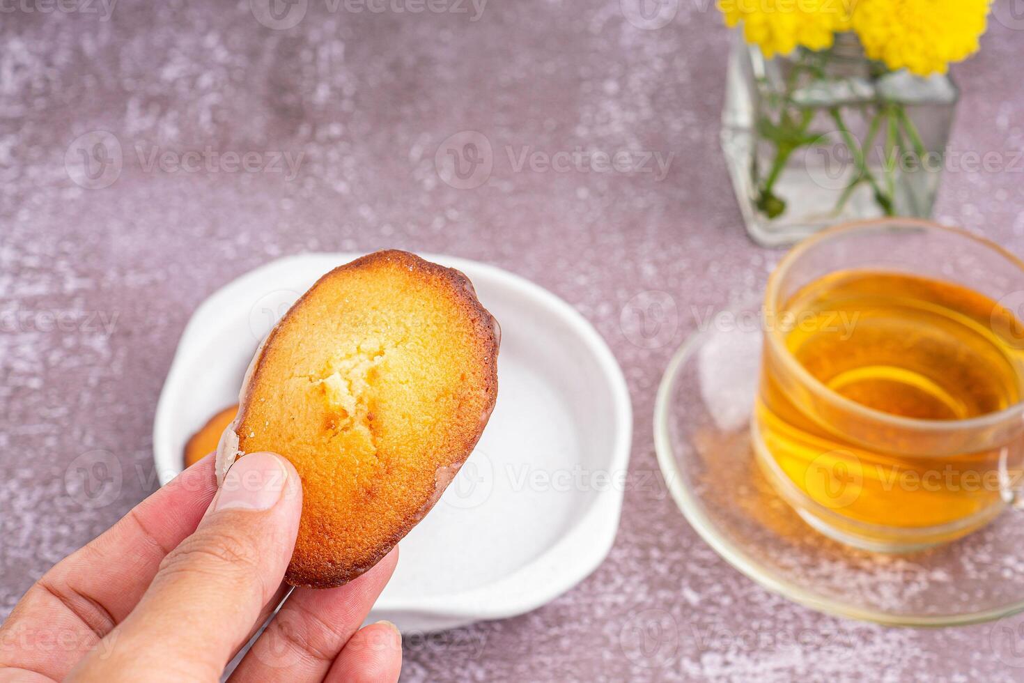 Hand holding a piece of madeleine on lemon glaze madeleines on a plate with a cup of tea placed on a gray stone background. Madeleine - homemade traditional French small cookie photo