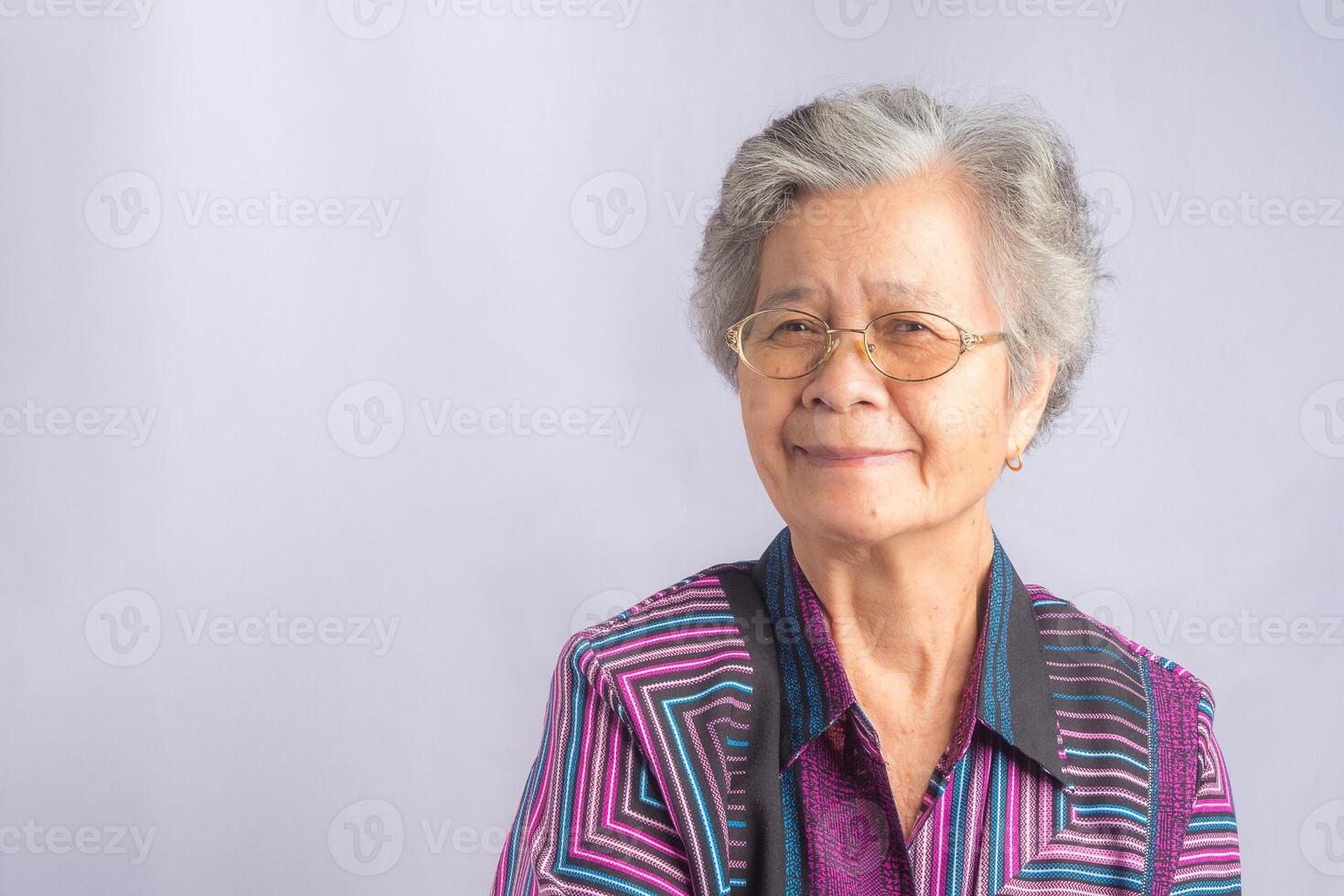 Portrait of a beautiful senior Asian woman wearing glasses with short white hair smiling and looking at the camera while standing with a gray background. Concept of aged people and healthcare photo