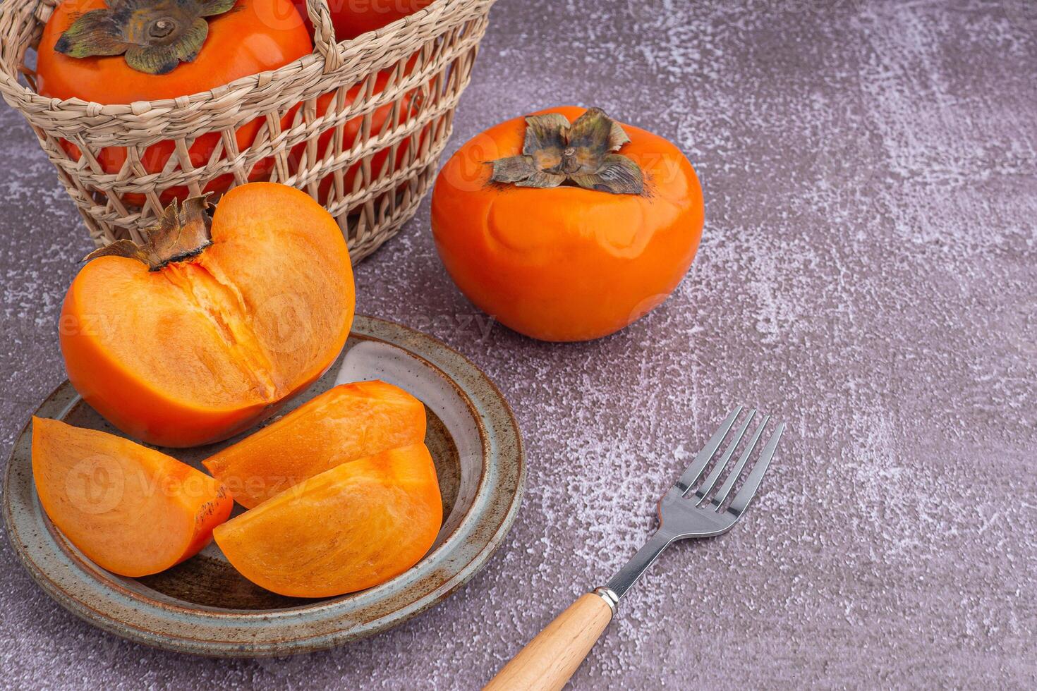Fresh ripe persimmons with slices of persimmon on a plate with cutlery and whole fruit in a basket with a white brick wall. Space for text. Close-up photo. Concept of health fruit photo