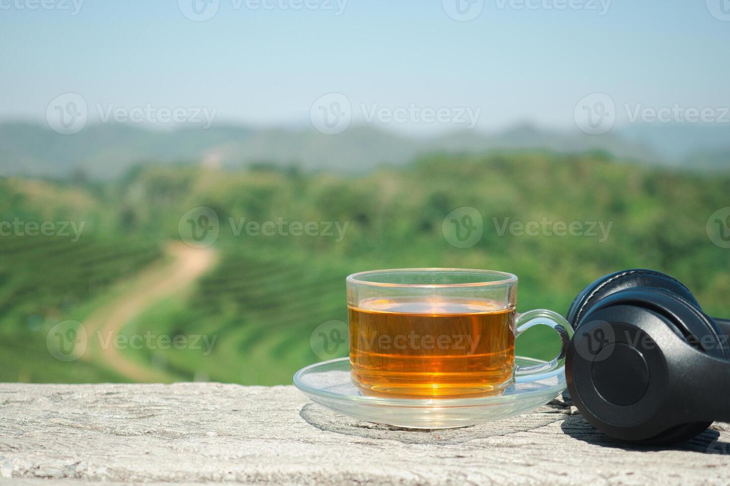 A cup of hot tea and headphones placed on the wooden table with the tea plantation background. Space for text. Close-up photo. Beautiful nature photo