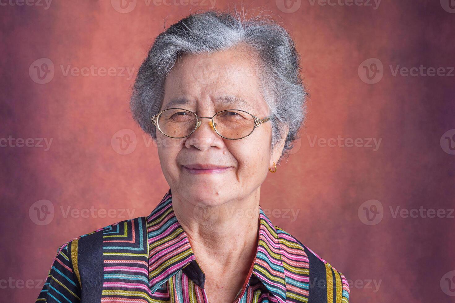 Portrait of a beautiful senior Asian woman wearing glasses with short white hair smiling and looking at the camera while standing with a vintage background. Concept of aged people and healthcare photo