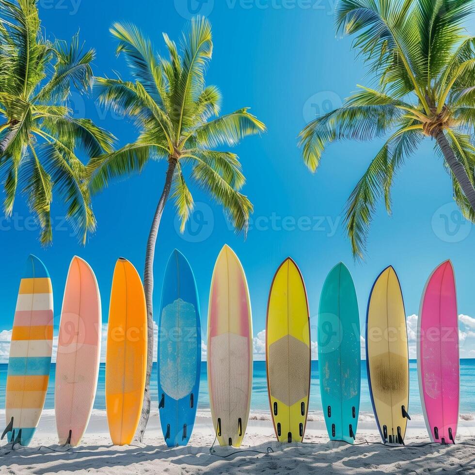 A collection of colorful surfboards lined up on a white sandy tropical beach surrounded by palm trees and the turquoise ocean in the background. photo