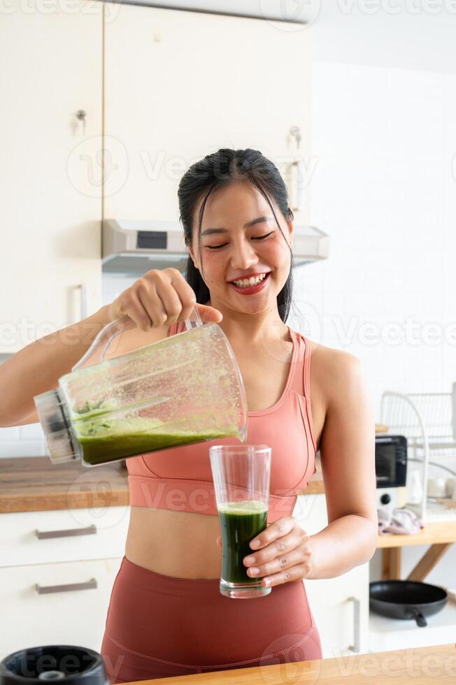 An attractive, healthy Asian woman in sportswear is pouring her green juice from a blender. photo