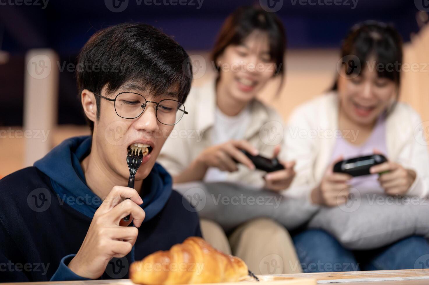 A young, happy Asian man is enjoying eating a croissant while his friends are playing a game. photo