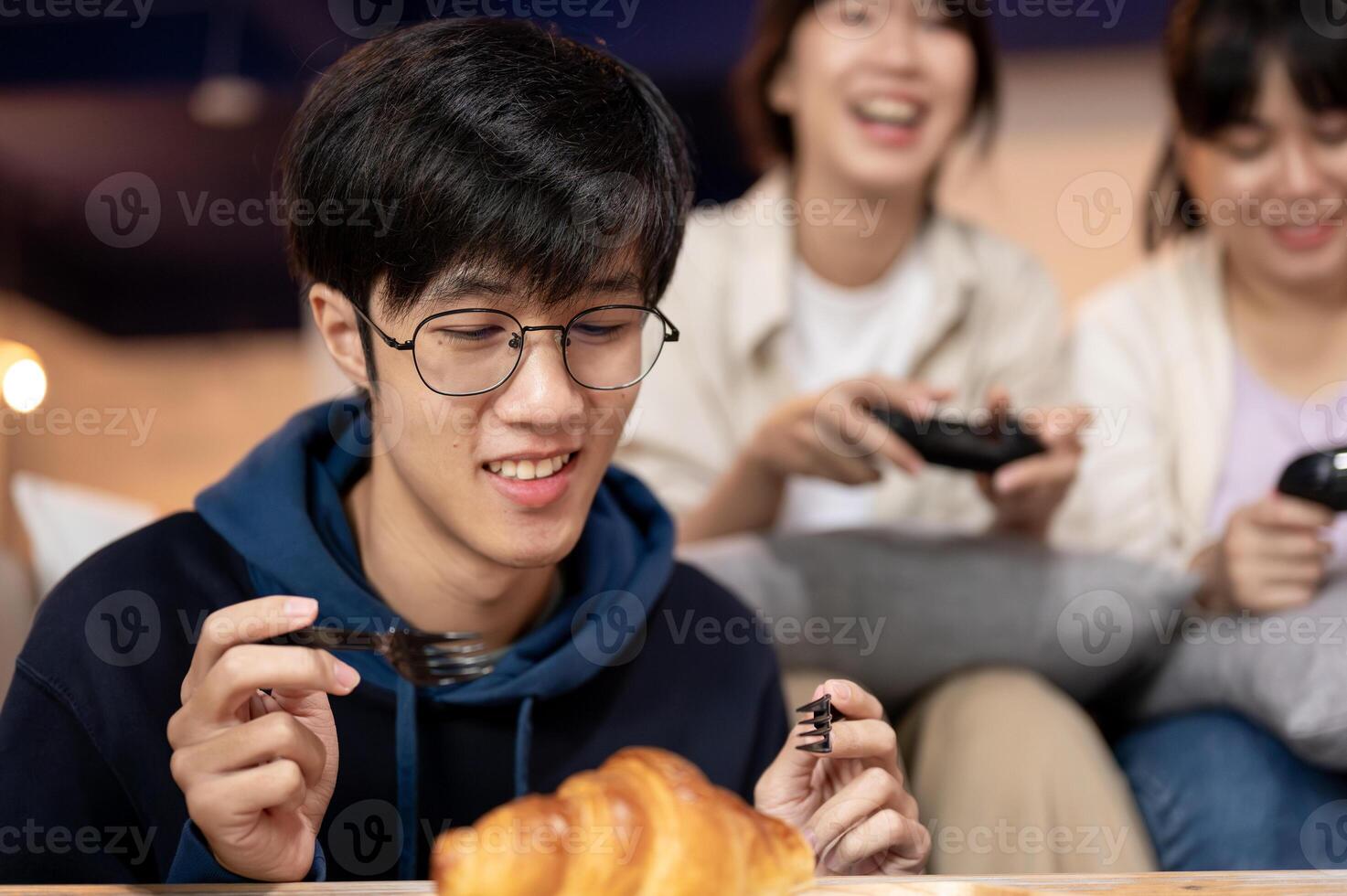 A young, happy Asian man is enjoying eating a croissant while his friends are playing a game. photo