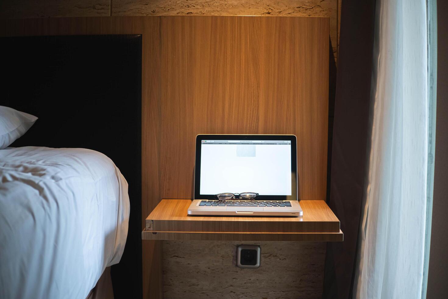 Laptop and glasses on a small desk next to a bed photo