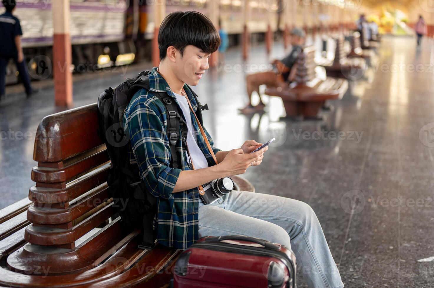 A man tourist is using his phone while sitting at a bench on a platform at a railway station. photo