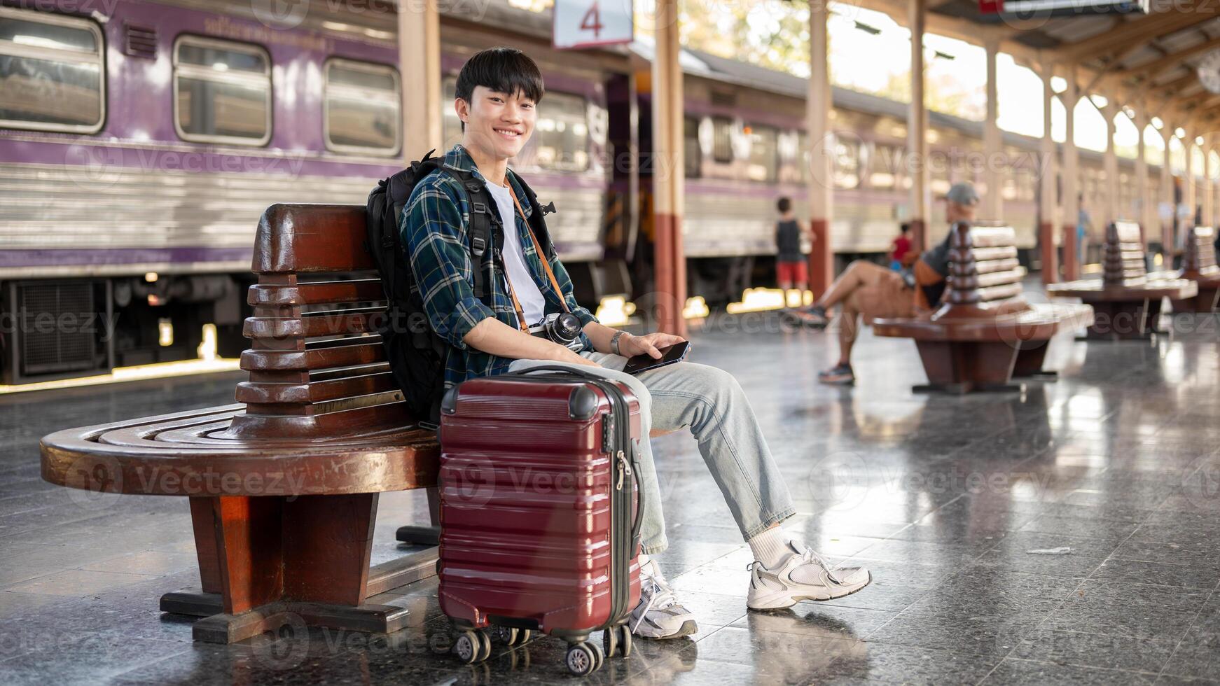 A happy Asian man sitting on a bench at a railway station with his bags, waiting for his train. photo