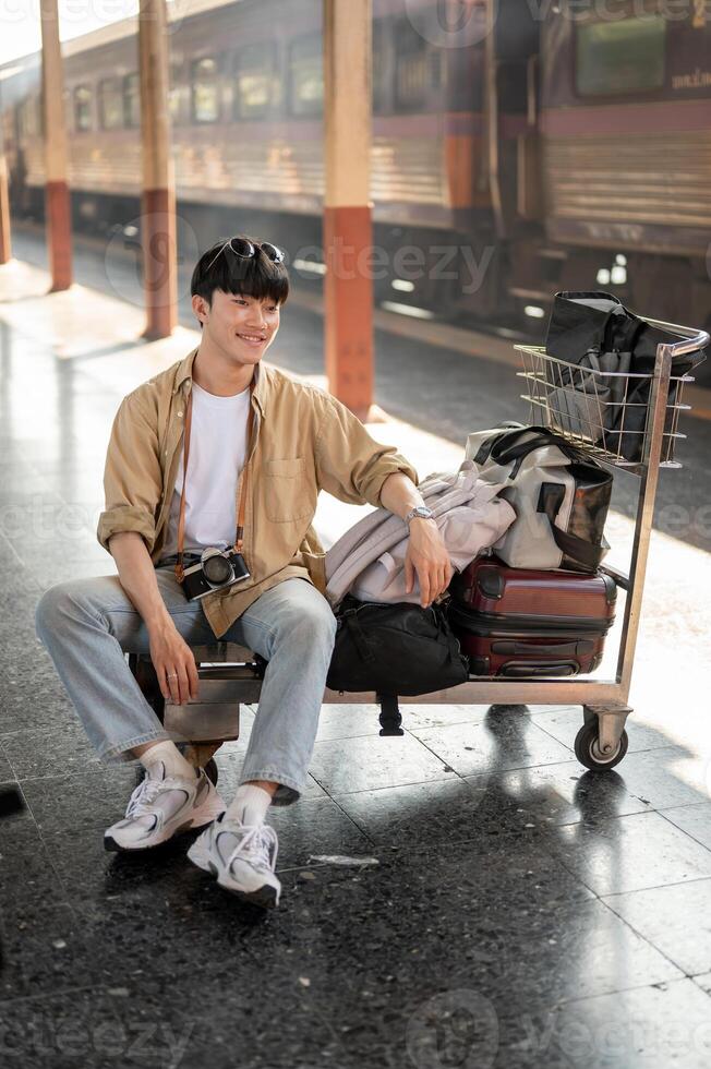 An Asian man traveler is sitting on a cart, waiting for his train at a railway station. photo