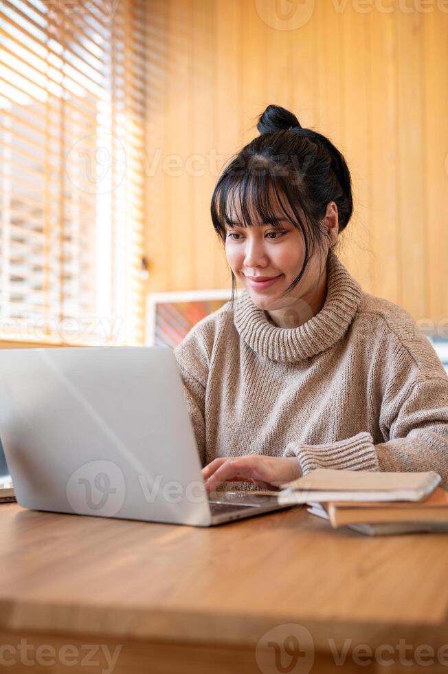 A positive Asian woman in a cosy sweater is working on her laptop computer in a cafe or library. photo