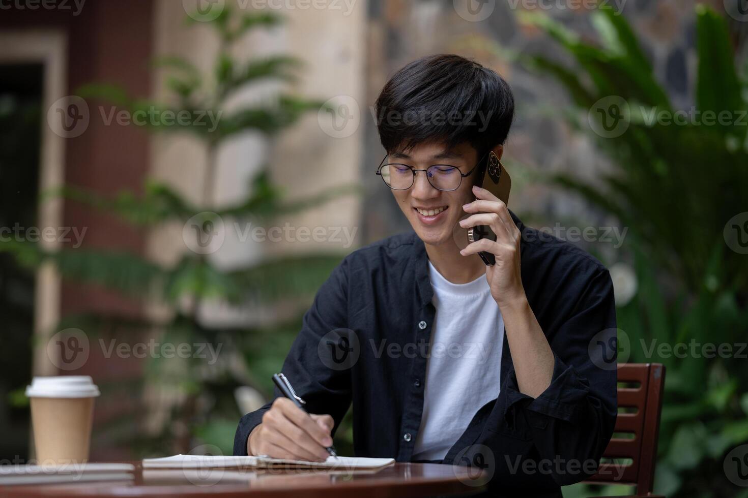 A happy Asian man is taking notes in his book while talking on the phone with someone outdoors. photo