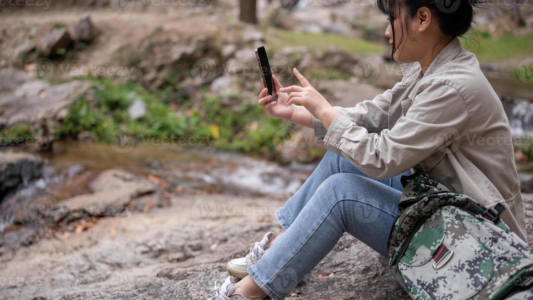 A female trekker is sitting on a rock and using her phone, taking a break while hiking. photo