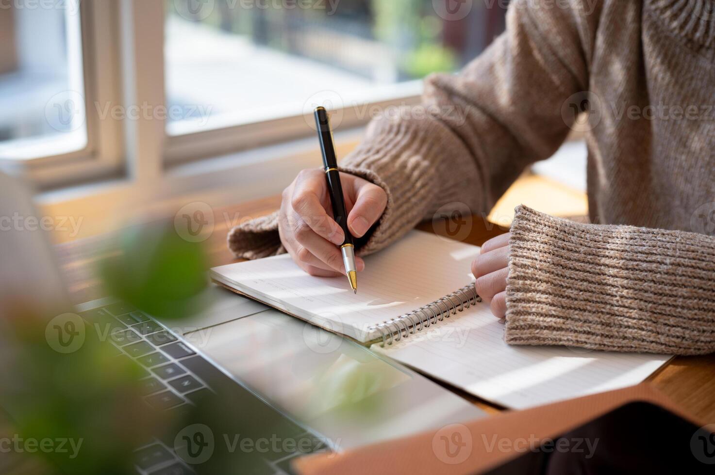 Close-up image of a woman is writing something in her notebook and working on her laptop indoors. photo