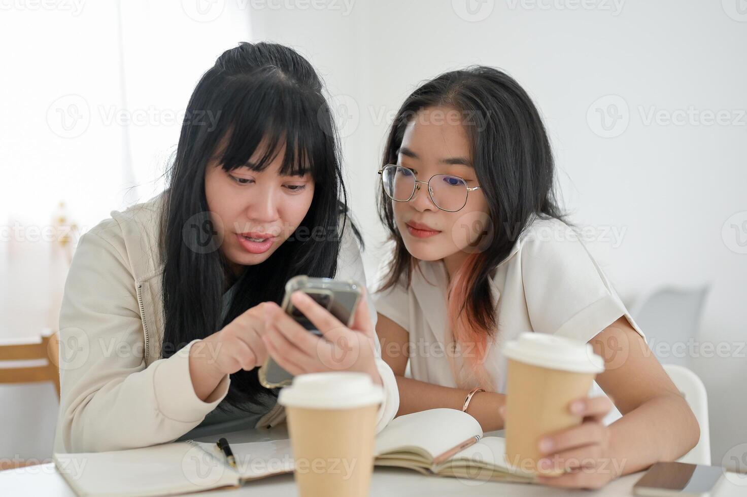 Two Asian female friends are watching something on a smartphone together while sitting in a cafe. photo