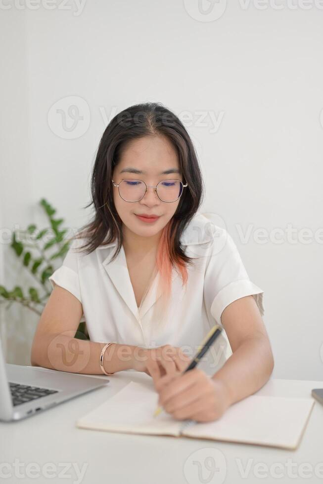 A focused Asian woman is working remotely from a cafe, taking notes or making list in her notebook photo