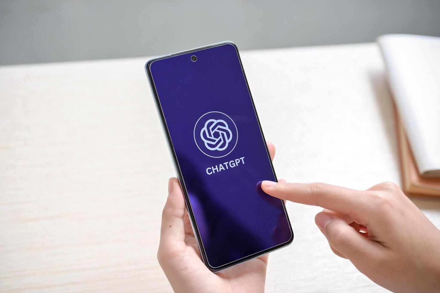 A woman's hand holding a smartphone with the ChatGPT logo on the screen at a table indoors. photo