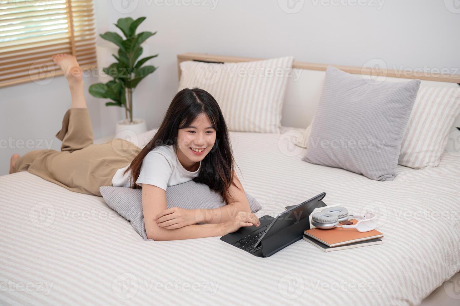 A Asian woman is lying on the bed and using her digital tablet, relaxing in her bedroom. photo
