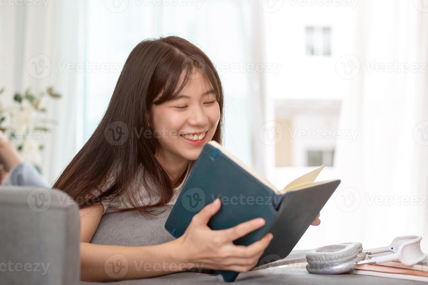 A young Asian woman is lying on a sofa, enjoying reading a book and relaxing in the living room. photo