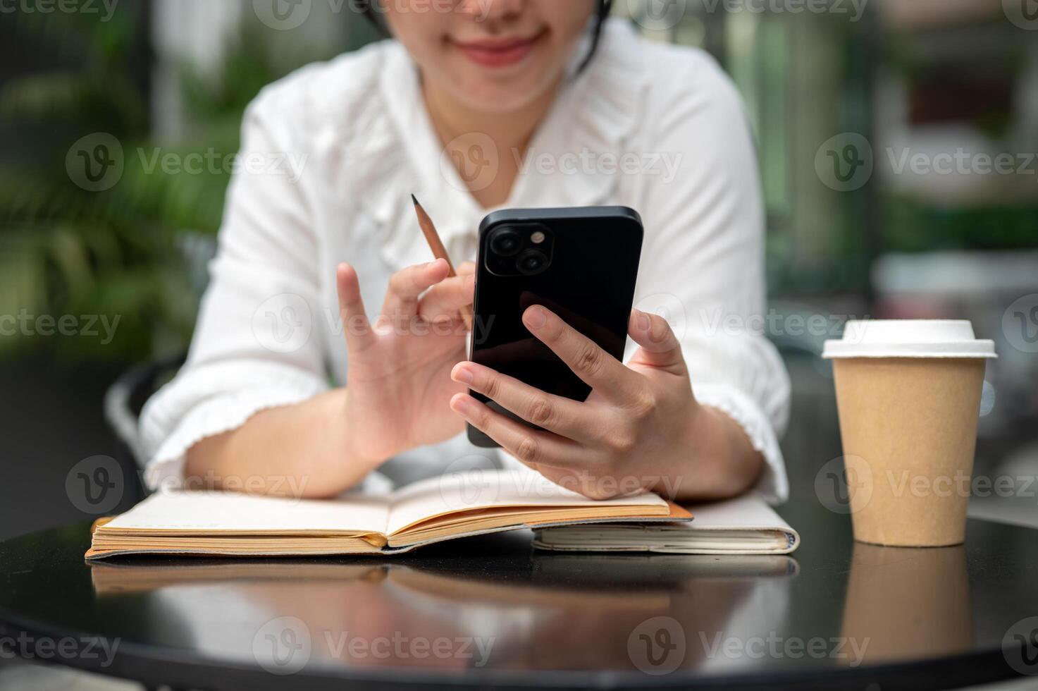 A woman using her smartphone while writing something in her notebook at a table. photo