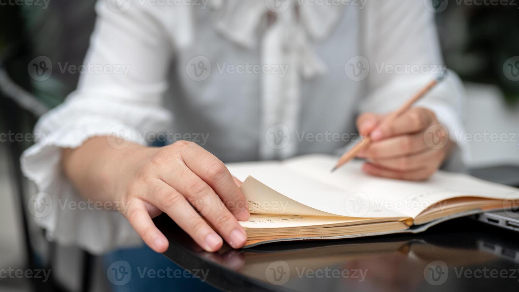 A close-up image of a woman sitting at a table, holding a pencil, and writing in a notebook. photo