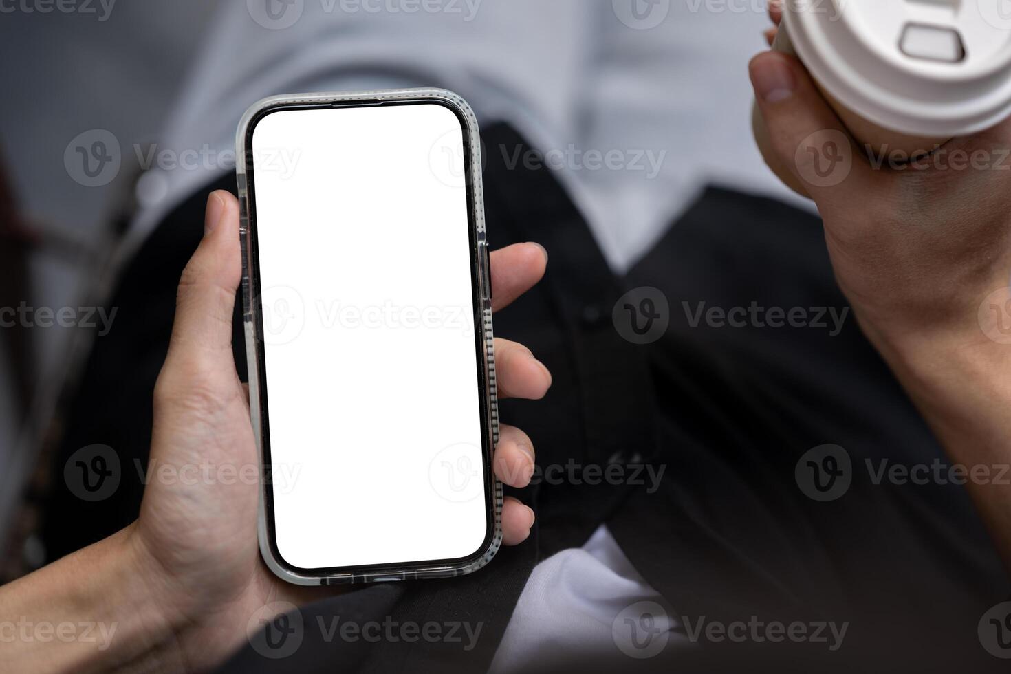 Close-up image of a man holding a white-screen smartphone and a coffee cup while sitting outdoors. photo