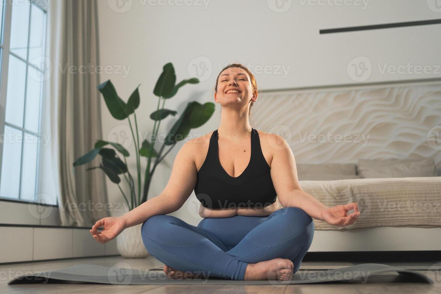 Yoga mindfulness meditation. Woman practicing yoga in living room at home. Plus size female sitting in lotus pose meditating smiling relaxing indoor. Girl doing breathing practice. Yoga at home photo