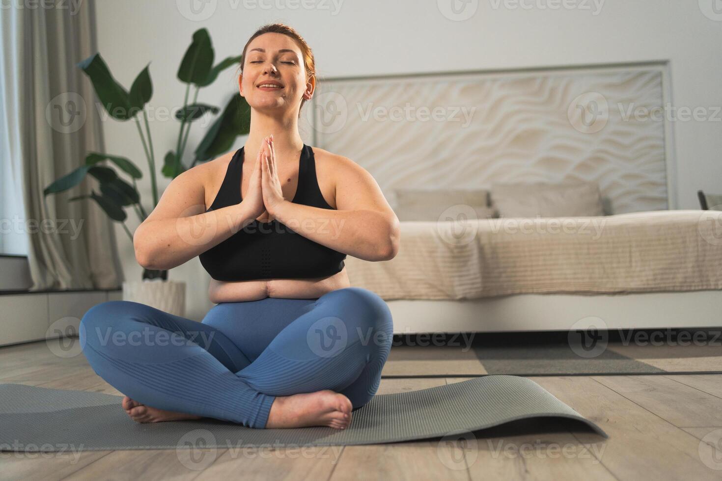 Yoga mindfulness meditation. Woman practicing yoga in living room at home. Plus size female sitting in lotus pose meditating smiling relaxing indoor. Girl doing breathing practice. Yoga at home photo