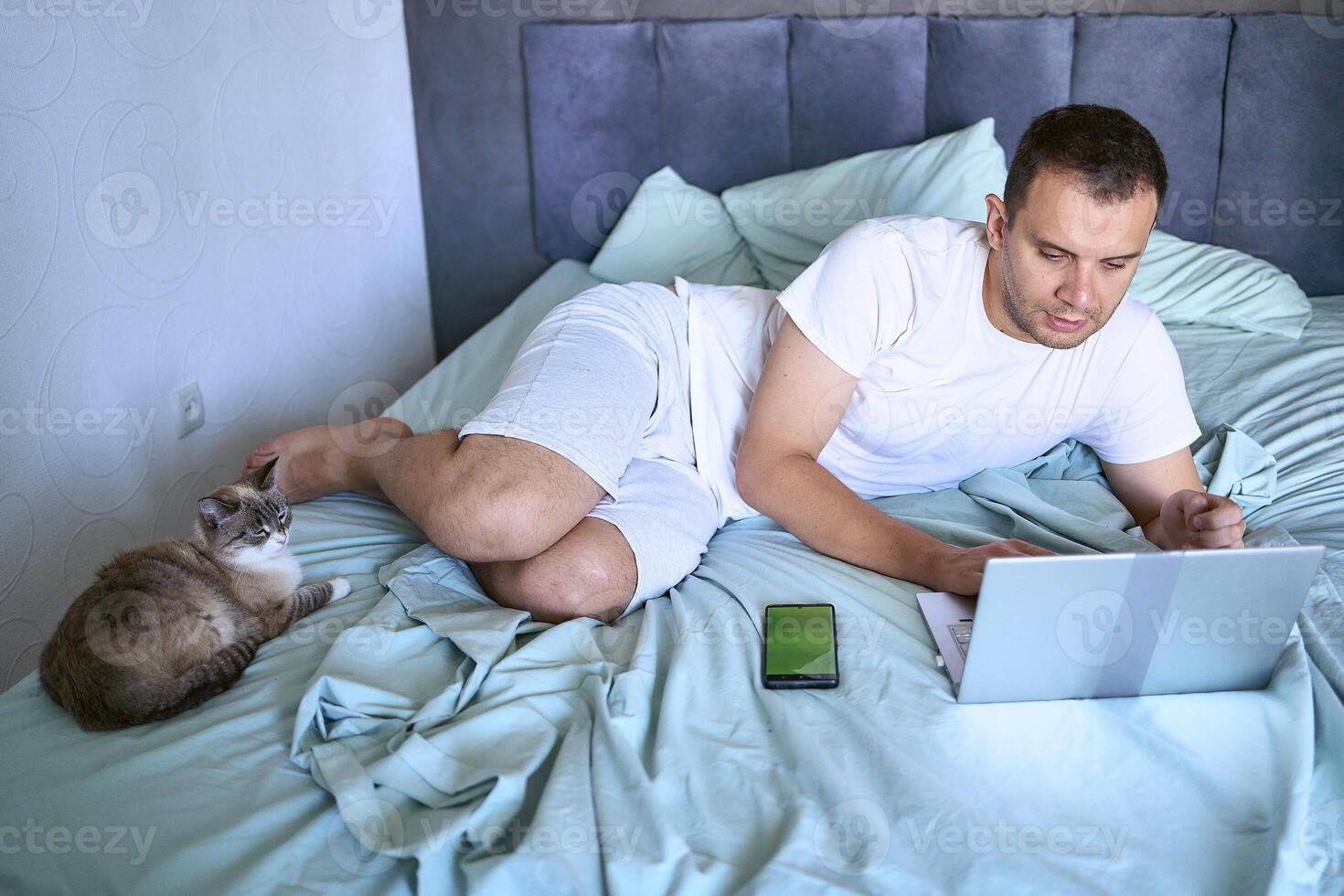 a young man is working on a laptop and a smartphone in bed, a cat is lying next to him photo