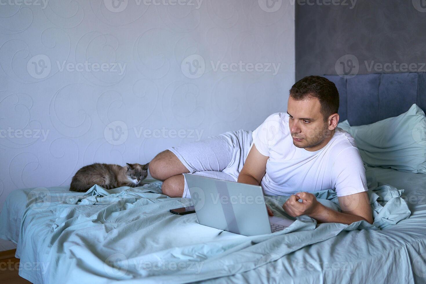 a young man is working on a laptop and a smartphone in bed, a cat is lying next to him photo