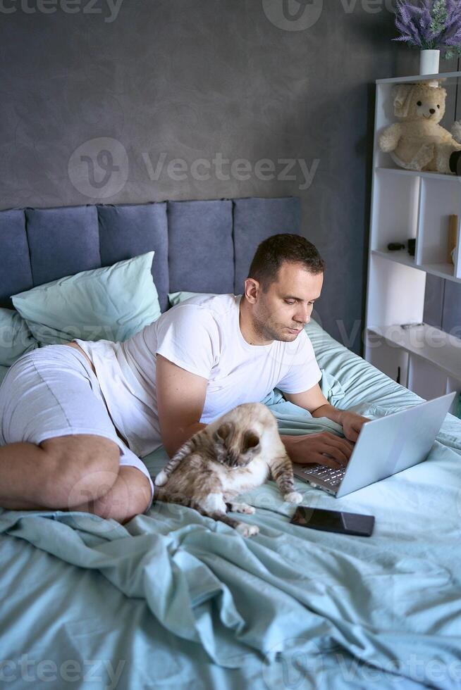 a young man is working on a laptop and a smartphone in bed, a cat is lying next to him photo