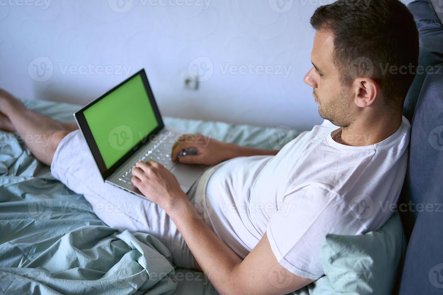 a young man is working on a laptop and a smartphone in bed, a cat is lying next to him photo