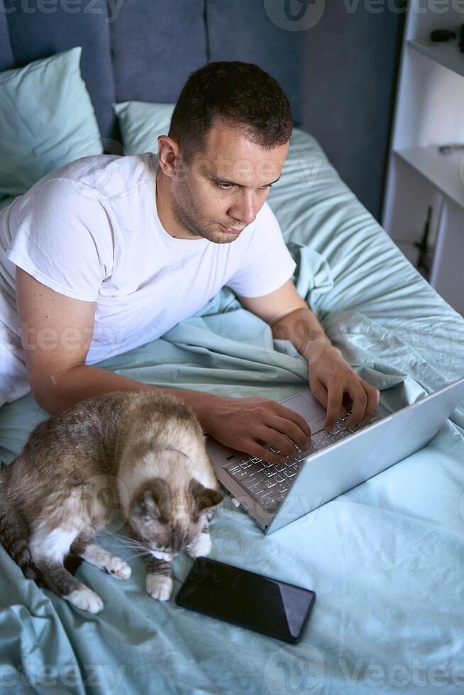 a young man is working on a laptop and a smartphone in bed, a cat is lying next to him photo