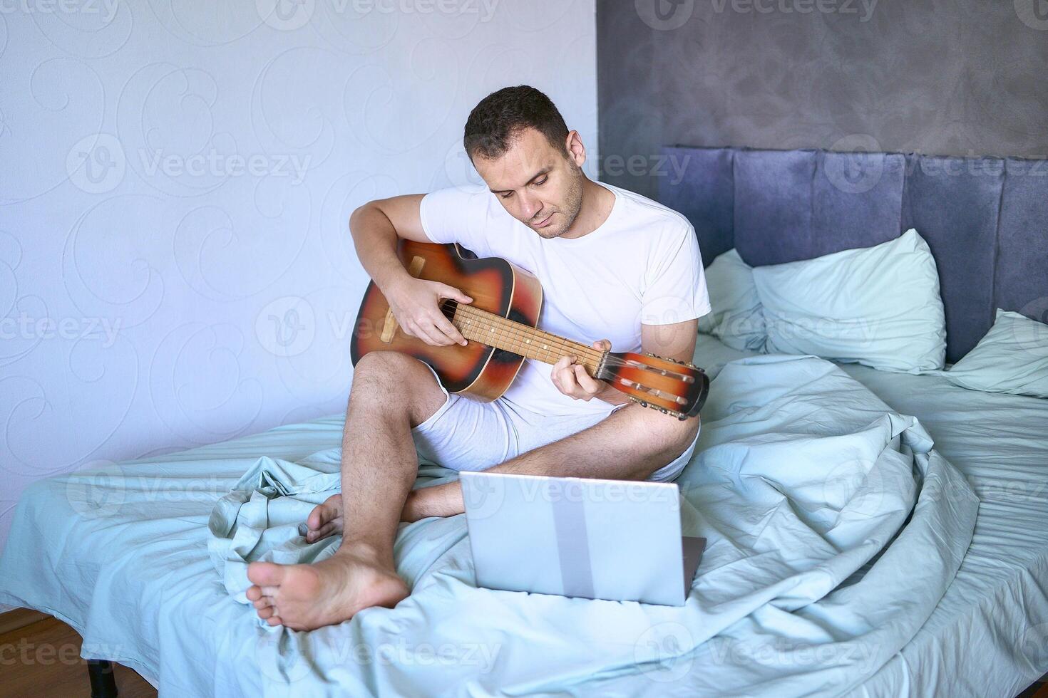 a young man is learning to play the guitar at home using online lessons on a laptop, the cat demands attention and interferes photo