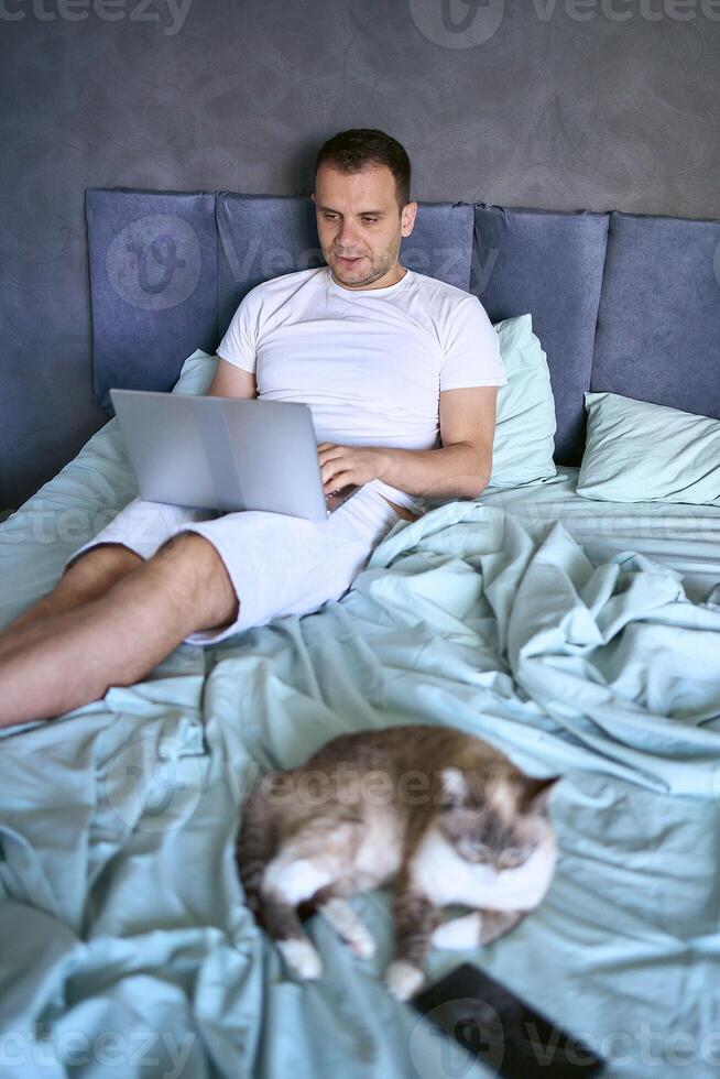 a young man is working on a laptop and a smartphone in bed, a cat is lying next to him photo
