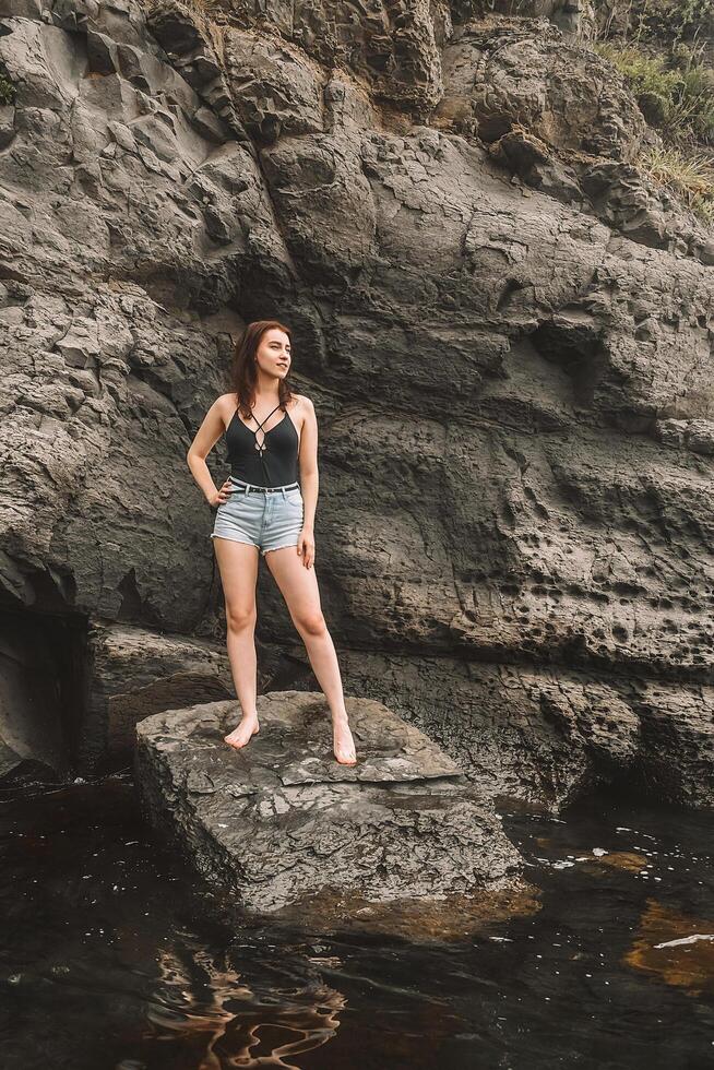A happy tourist in a swimsuit and sunglasses likes to take pictures in nature as a souvenir. A female traveler poses on a beach by the sea surrounded by volcanic rocks and shares the joy of travel photo