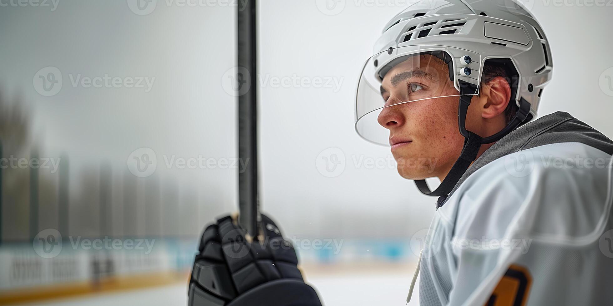 A hockey player in a white helmet and jersey stands sideways and holds a stick. Hockey arena in the background. Copy space photo