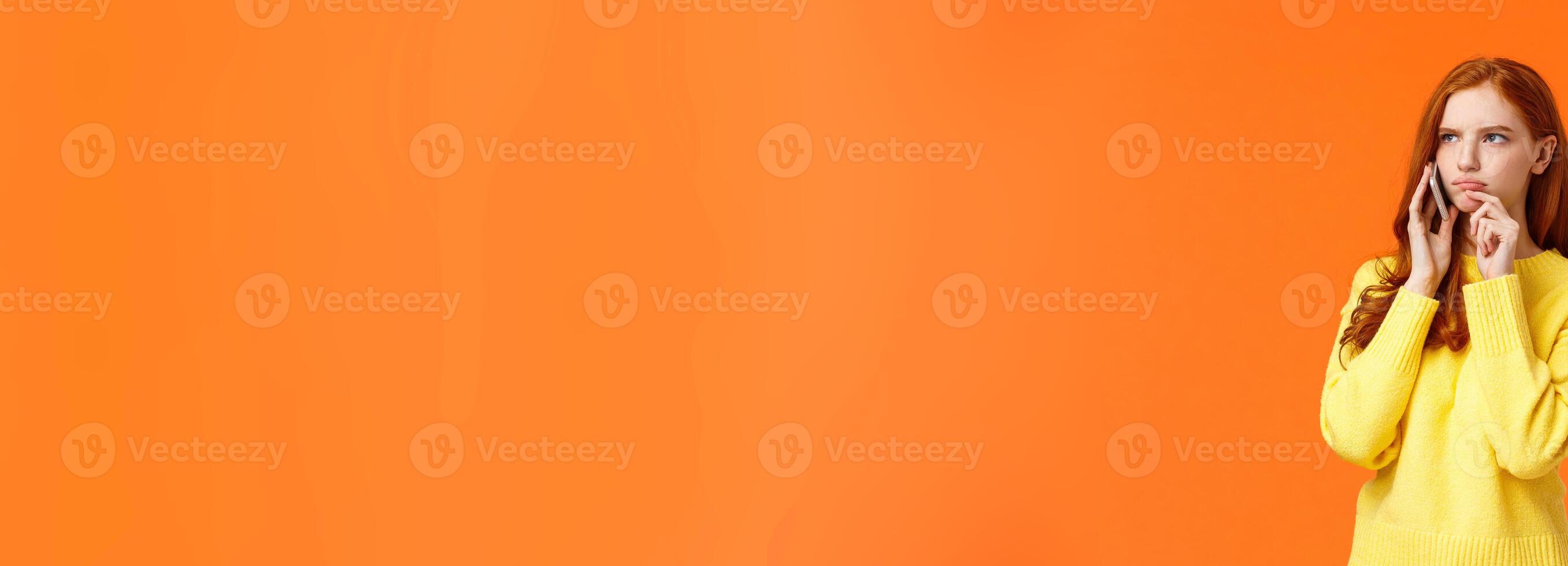 Indecisive and unsure, skeptical serious-looking redhead woman having tough decision make during conversation on phone, frowning touch lip pensive, order food delivery, orange background photo
