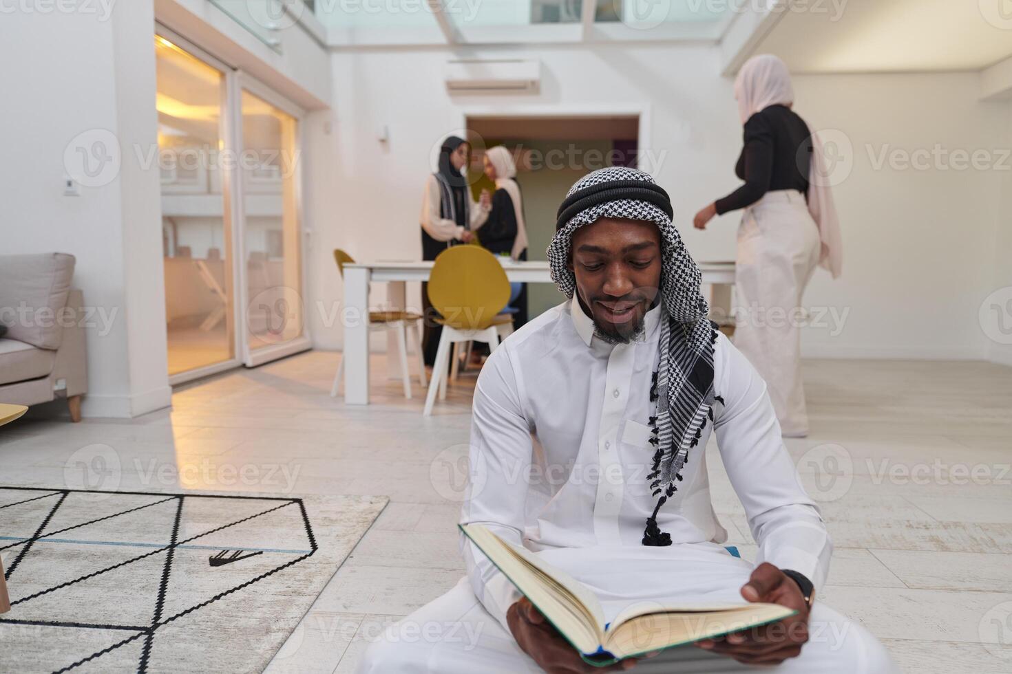 In the sacred month of Ramadan, an African American Muslim man engrossed in reading the Holy Quran is surrounded by a backdrop where three hijab-wearing women prepare and serve food for iftar photo