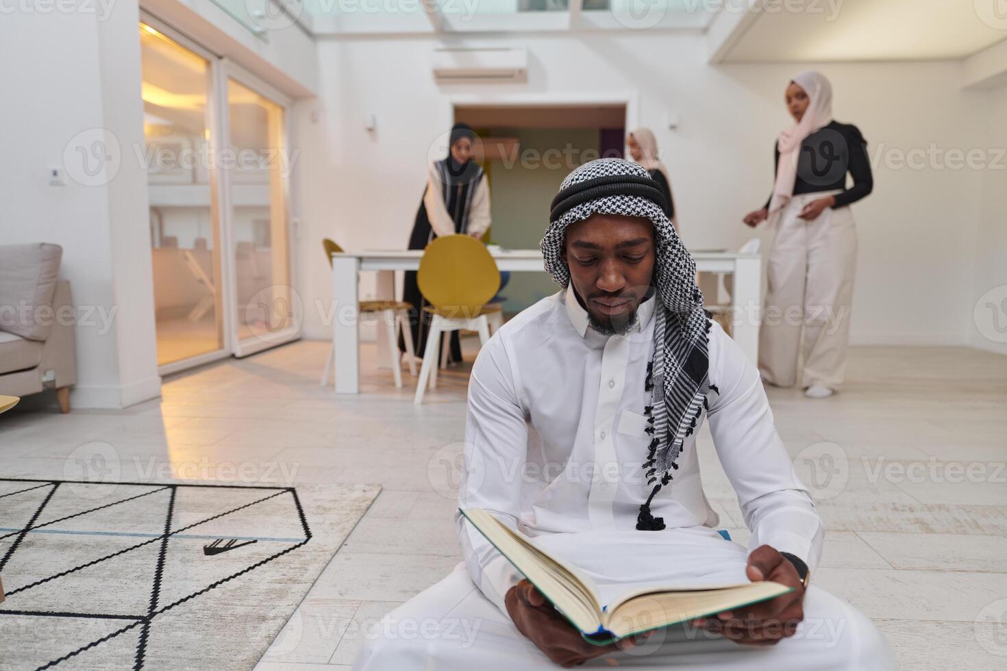 In the sacred month of Ramadan, an African American Muslim man engrossed in reading the Holy Quran is surrounded by a backdrop where three hijab-wearing women prepare and serve food for iftar photo